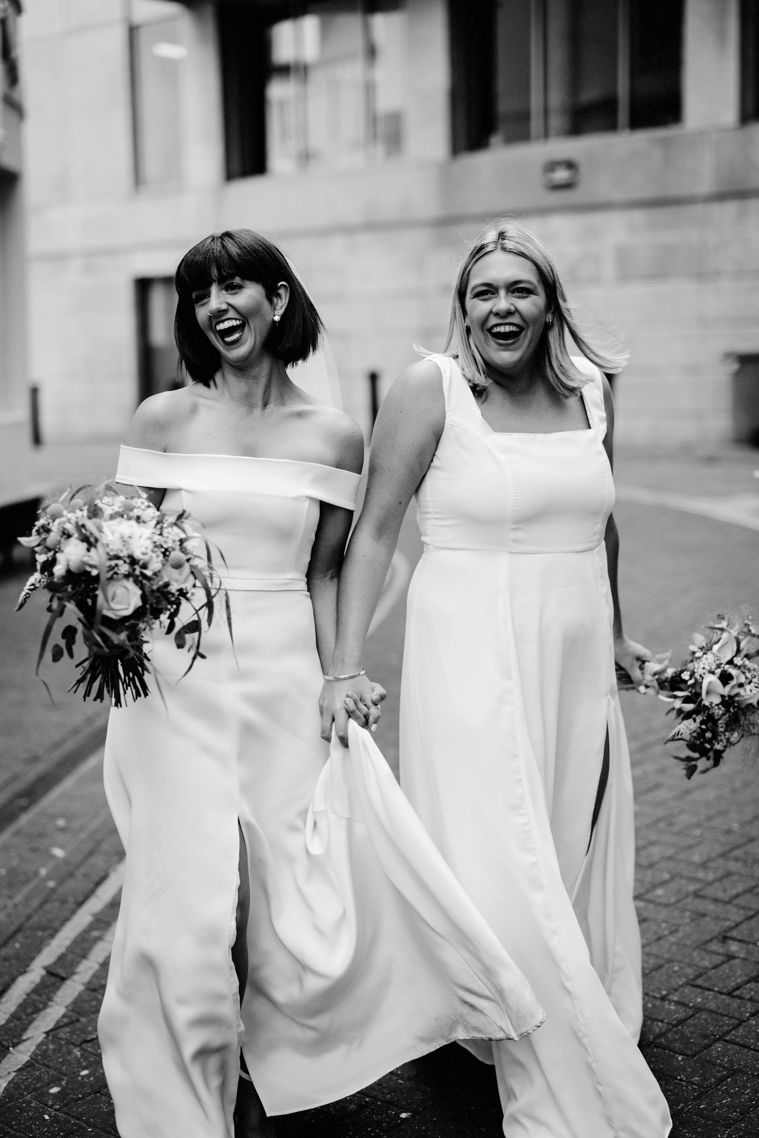 A joyful wedding photo captured by Teddy Photo + Film, showing two brides walking hand in hand through the streets in matching modern white dresses. Both women are laughing openly as they hold their bouquets and lift their skirts slightly while walki