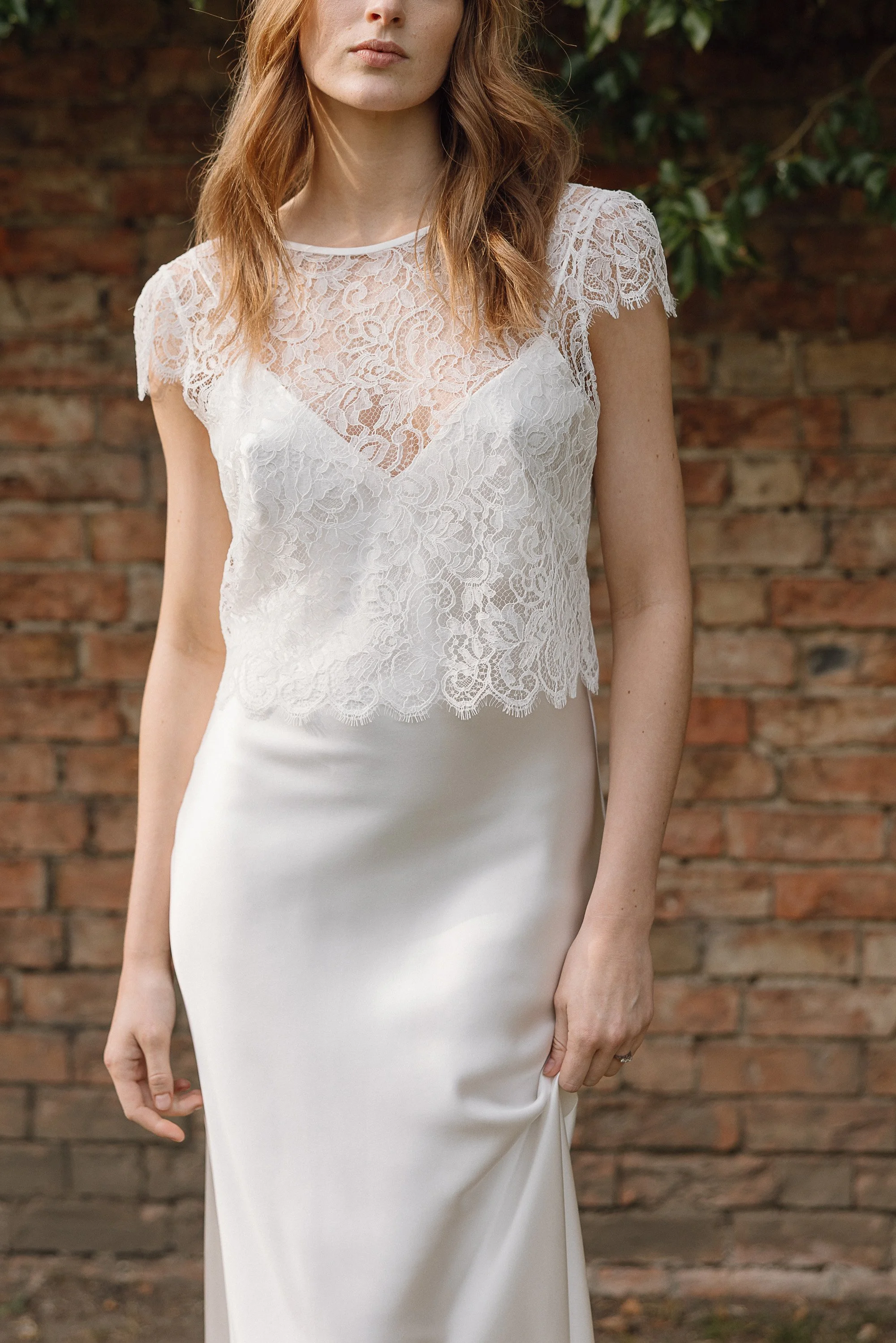 The bride stands before a warm brick wall wearing a delicate lace top layered over a smooth silk slip, the scalloped edges and intricate floral pattern catching the light while her relaxed posture and softly draped skirt create an effortlessly romant