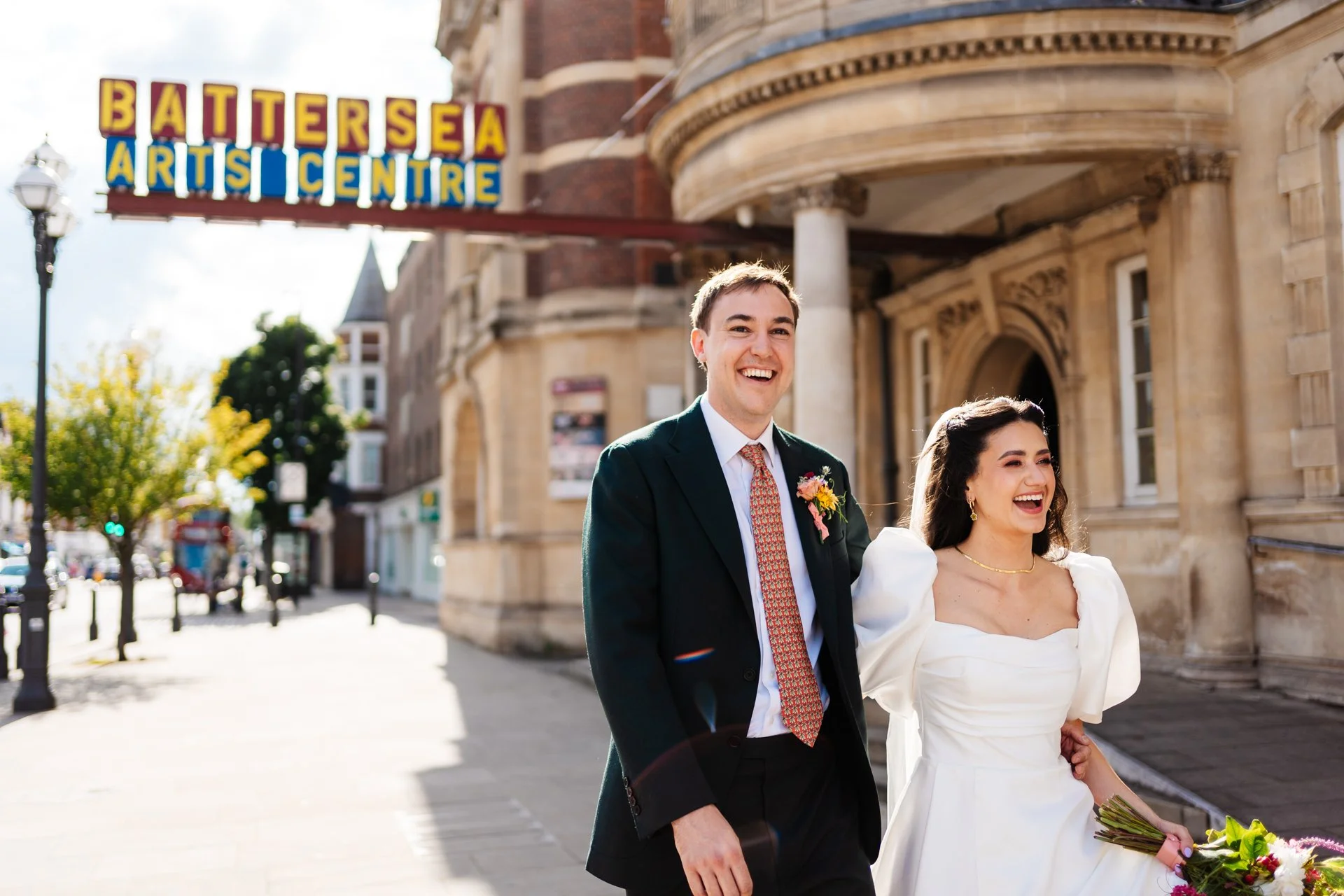A couple walks together outside the Battersea Arts Centre on a sunny day, smiling as they move along the pavement in front of the building’s curved stone façade. The person on the left wears a dark suit with a patterned tie and a floral boutonniere, 