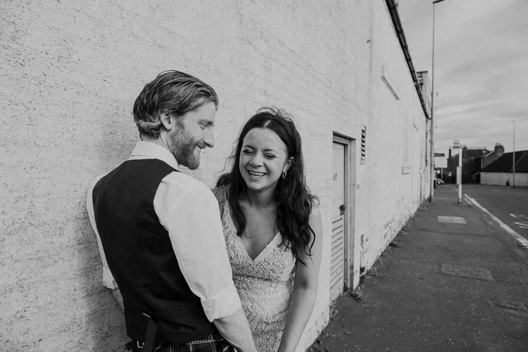 A relaxed, laughter-filled moment captured by Mirrorbox Photography, showing a couple standing together against a textured white wall on a quiet suburban street. The bride’s sparkly, modern dress catches the light, while the groom—dressed casually in