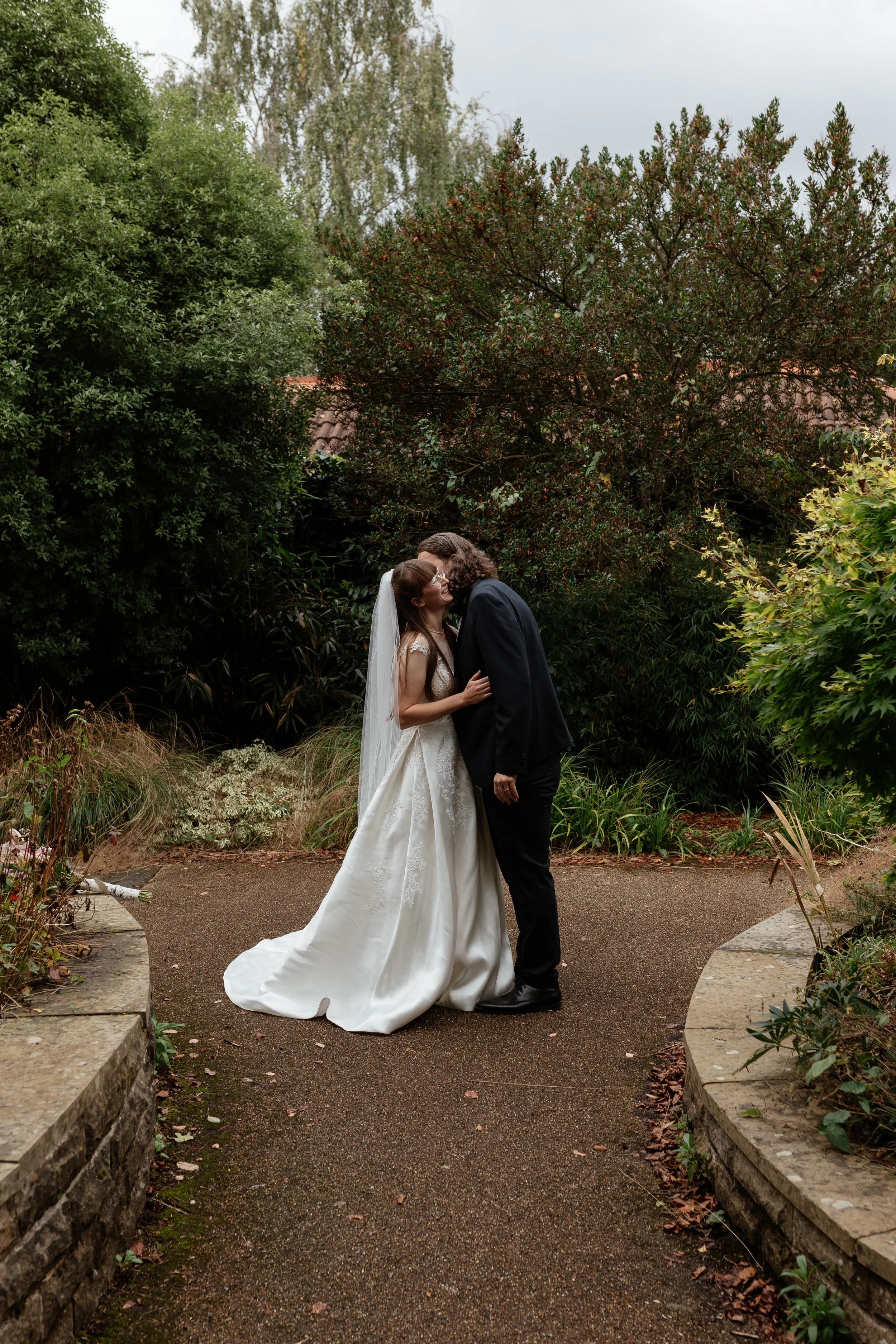 A romantic garden wedding portrait captured by Laura Wilson Photography, showing a couple sharing a gentle kiss along a winding path surrounded by lush greenery. The bride wears a flowing satin gown with a long veil that drapes softly behind her whil