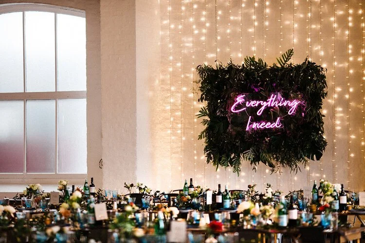Modern wedding reception with a striking greenery installation by Catherine Short Florals featuring lush foliage surrounding a neon sign that reads Everything I Need. Styled against a backdrop of warm fairy lights with colourful table arrangements be