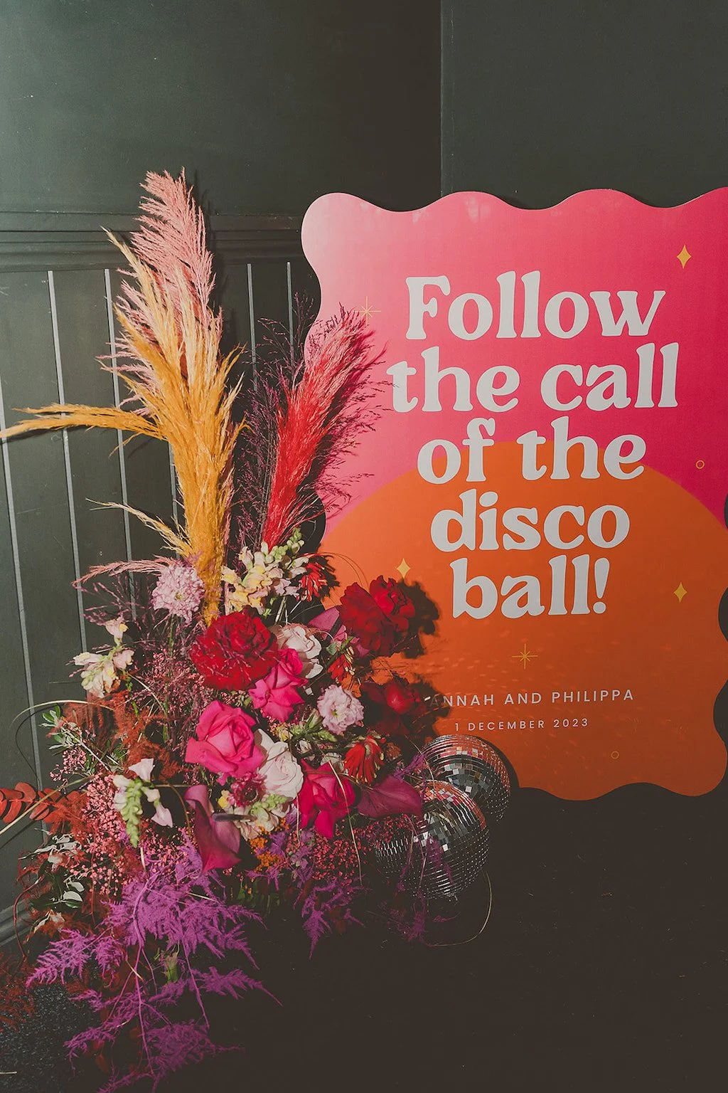 Vibrant wedding signage by AMW Events reading 'Follow the call of the disco ball!' with bold florals, pampas grass, and a disco ball accent. Laura Rhian Photography.