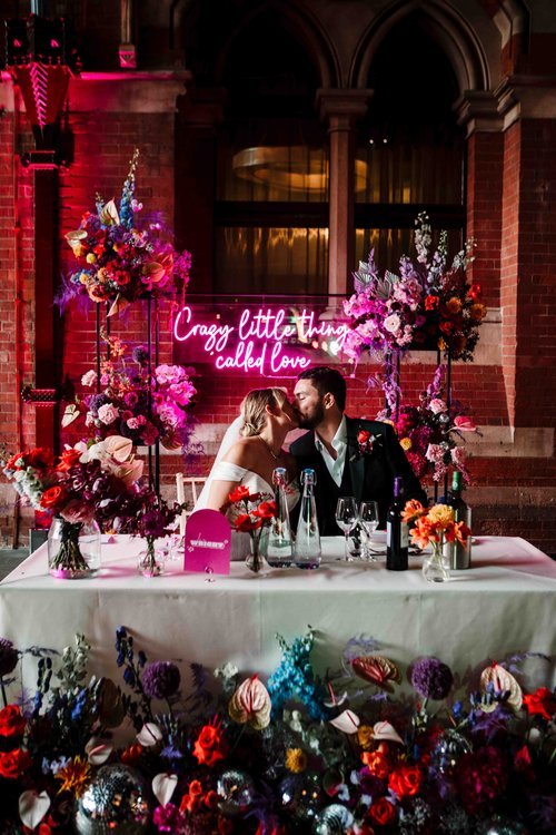 A vibrant and artistic wedding reception portrait captured by Amy Faith Photography, showing the couple sharing a celebratory kiss behind their beautifully decorated sweetheart table. Rich, saturated tones from the floral arrangements and neon sign c