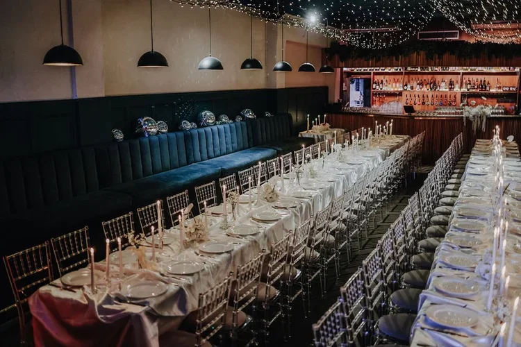 An elegant wedding reception setup inside Colours Hoxton in Shoreditch, London. The room features long banquet-style tables dressed in white linens, soft candlelight, and clear ghost chairs, creating a chic modern atmosphere. Warm pendant lighting an