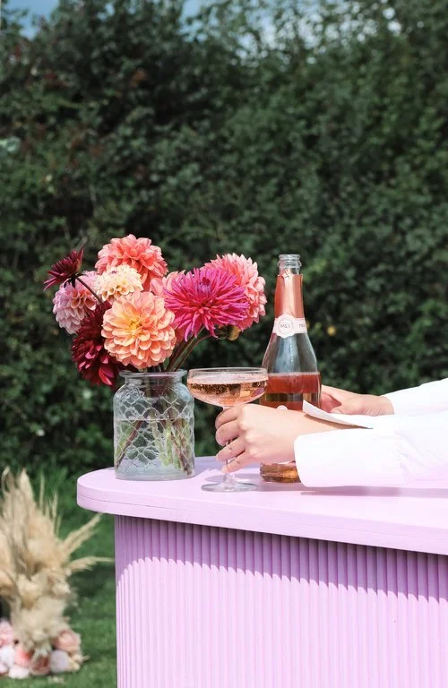 A stylish pink bar setup by The Pink Drinks Van featuring a glass of sparkling rosé beside a bottle of fizz and a vase filled with vibrant dahlias. Set outdoors against lush greenery, this elegant scene captures the brand’s signature blend of luxury,