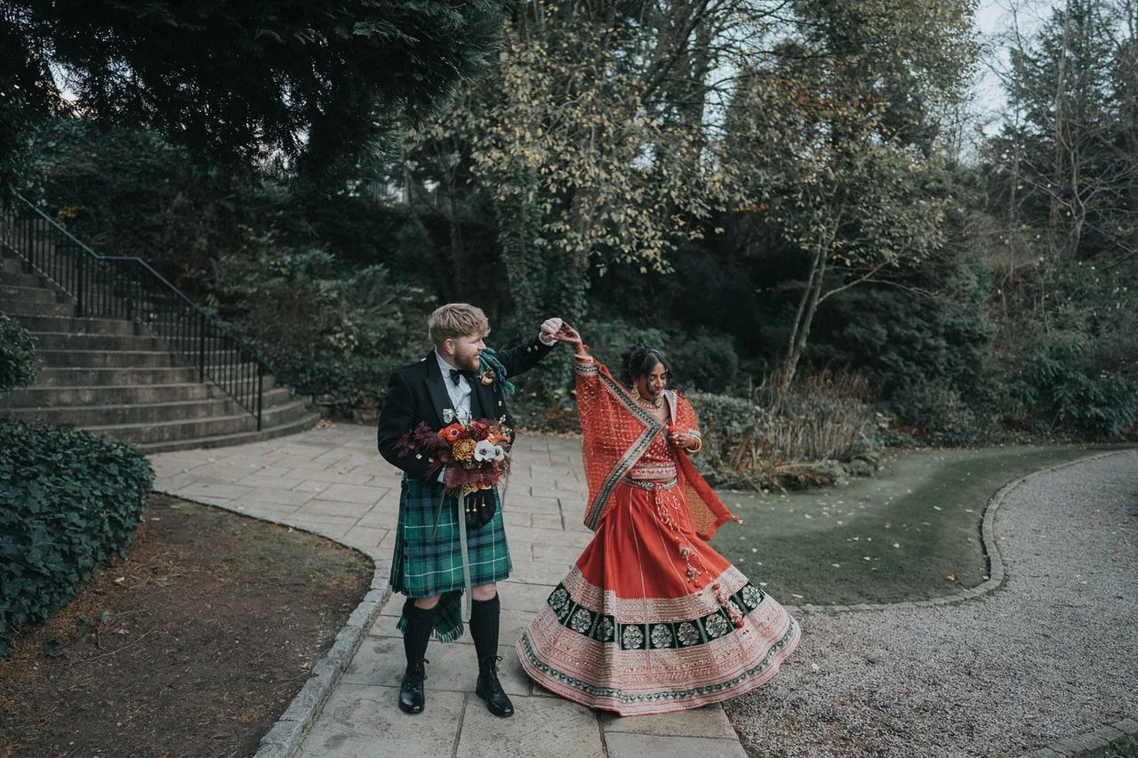 A couple dance together on a quiet garden path, captured mid-twirl. One partner wears a traditional green tartan kilt with a dark jacket and holds a rich, autumnal bouquet, while the other spins gracefully in a vibrant red and gold embroidered dress 