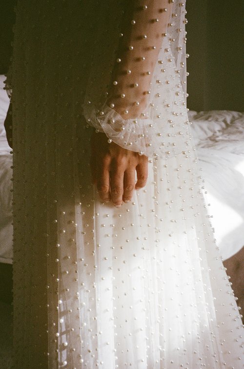 A close up, film shot captures the delicate detail of a bride’s pearl adorned gown as soft light falls across the sheer fabric. The gentle texture of the beading and the relaxed position of her hand create a quiet, intimate moment in the calm before 