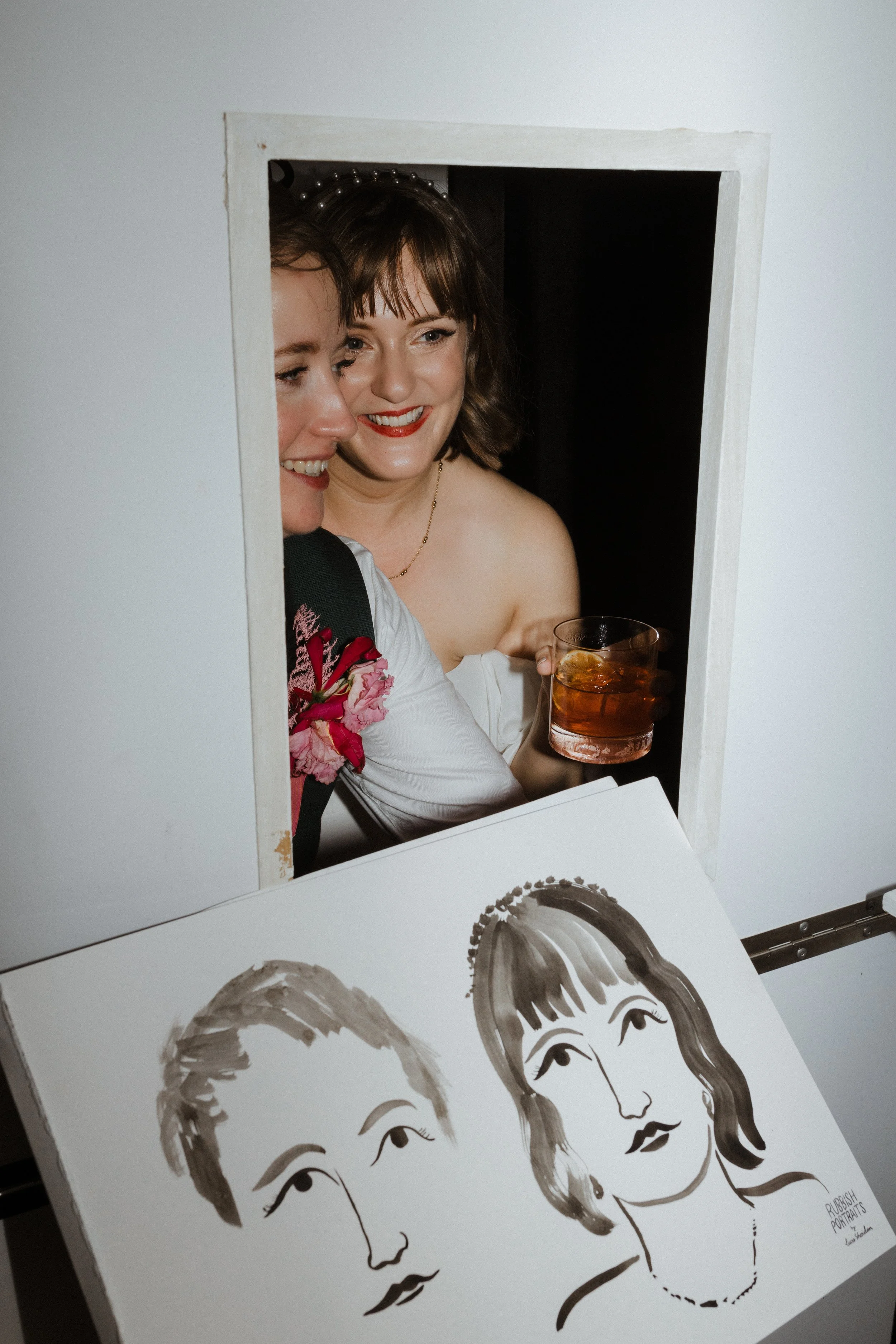 Two newlyweds smiling inside the ‘Rubbish Portraits’ live art booth by Lucie Sheridan, holding drinks and admiring their hand-drawn caricature portraits. The playful black-and-white artwork captures their likeness with a whimsical touch, perfectly re