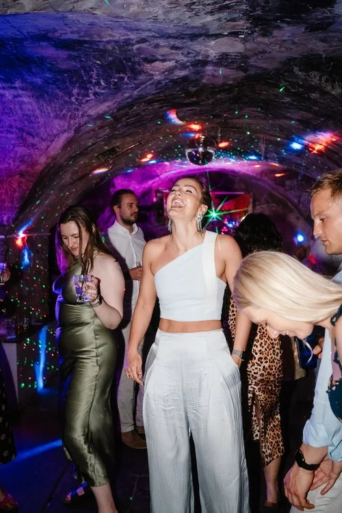 A guest throws her head back in laughter on a lively underground dance floor filled with colourful lights, laser patterns, and the energy of a packed celebration. Dressed in a modern one-shoulder top and wide-leg trousers, she moves confidently among