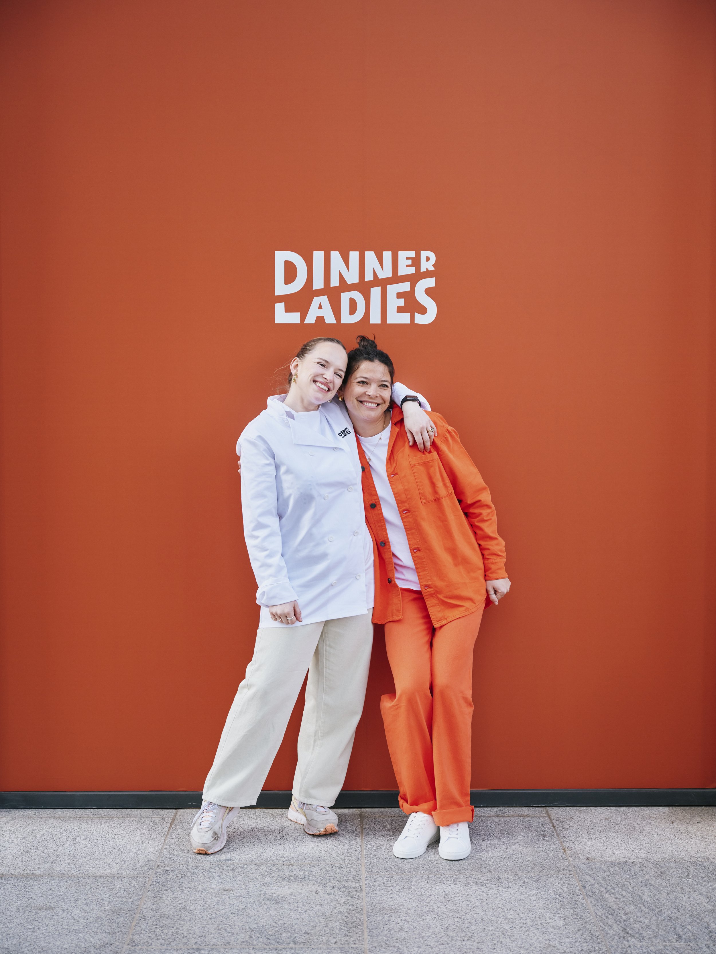 Dinner Ladies founders, Emily and Lily. They are standing in front of an orange background and giving off best mate energy.