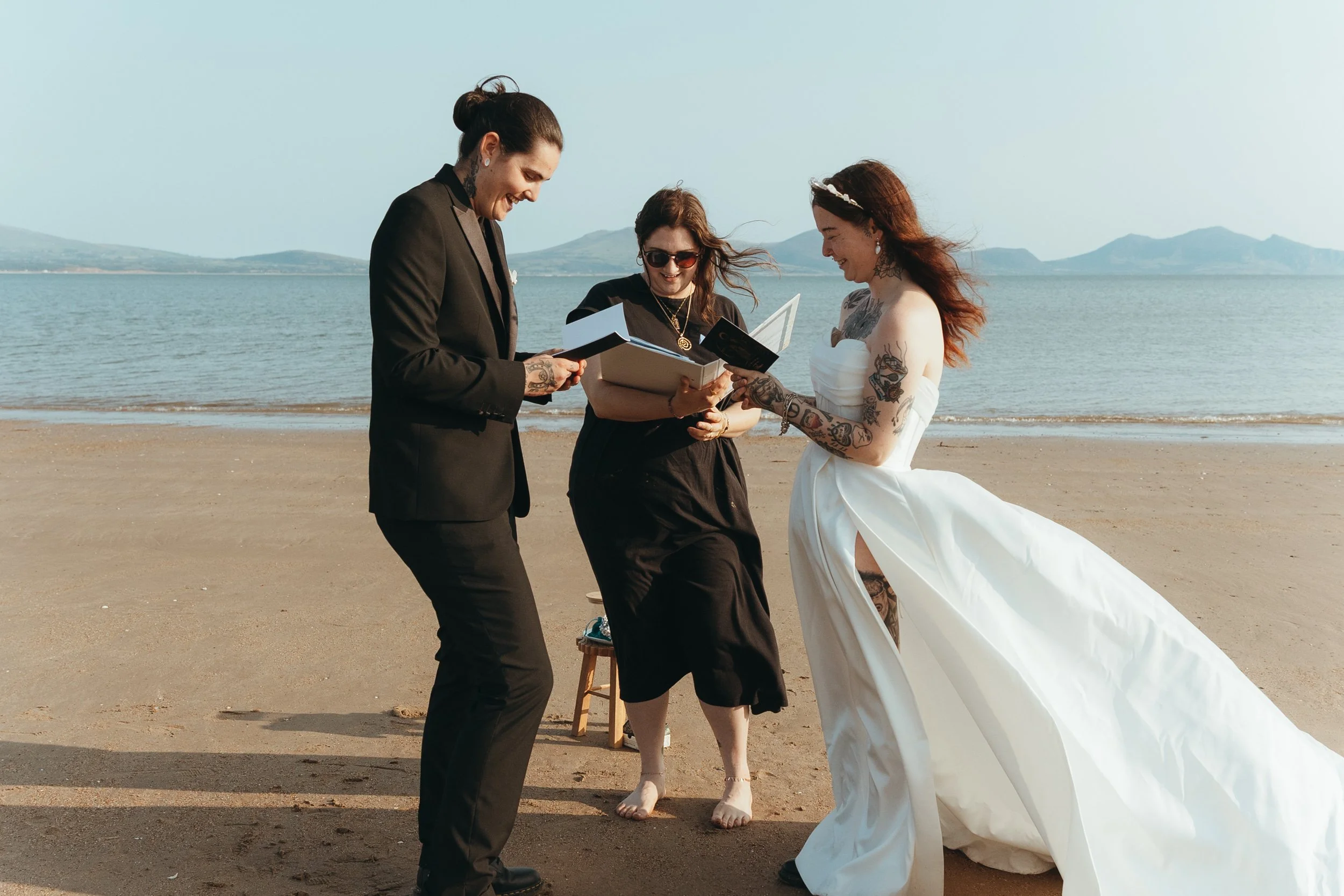 This image captures a joyful beach wedding ceremony. A couple - one wearing a black suit and the other a strapless white gown - are standing barefoot on the sand, laughing and exchanging vows. Between them stands Lauren The Spiritual Celebrant dresse