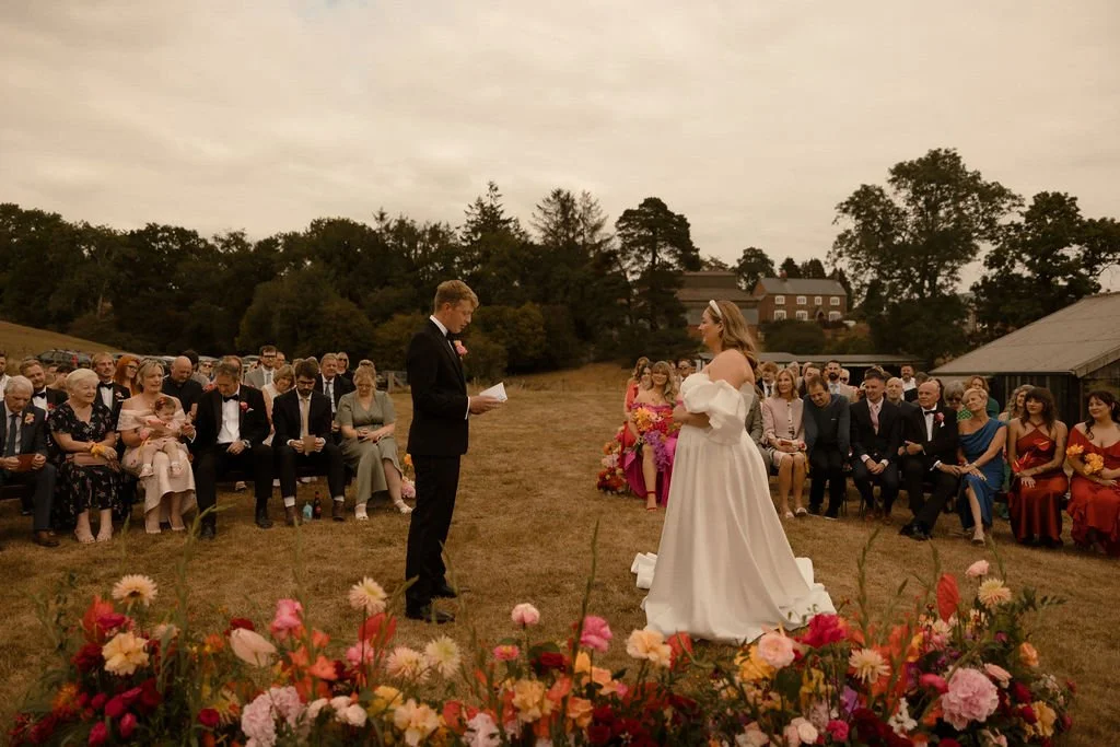A romantic outdoor wedding ceremony at The Giraffe Shed in Wales, where the couple exchange heartfelt vows surrounded by family and friends. The bride stuns in an elegant off-the-shoulder white gown, while the groom reads his vows against a scenic co