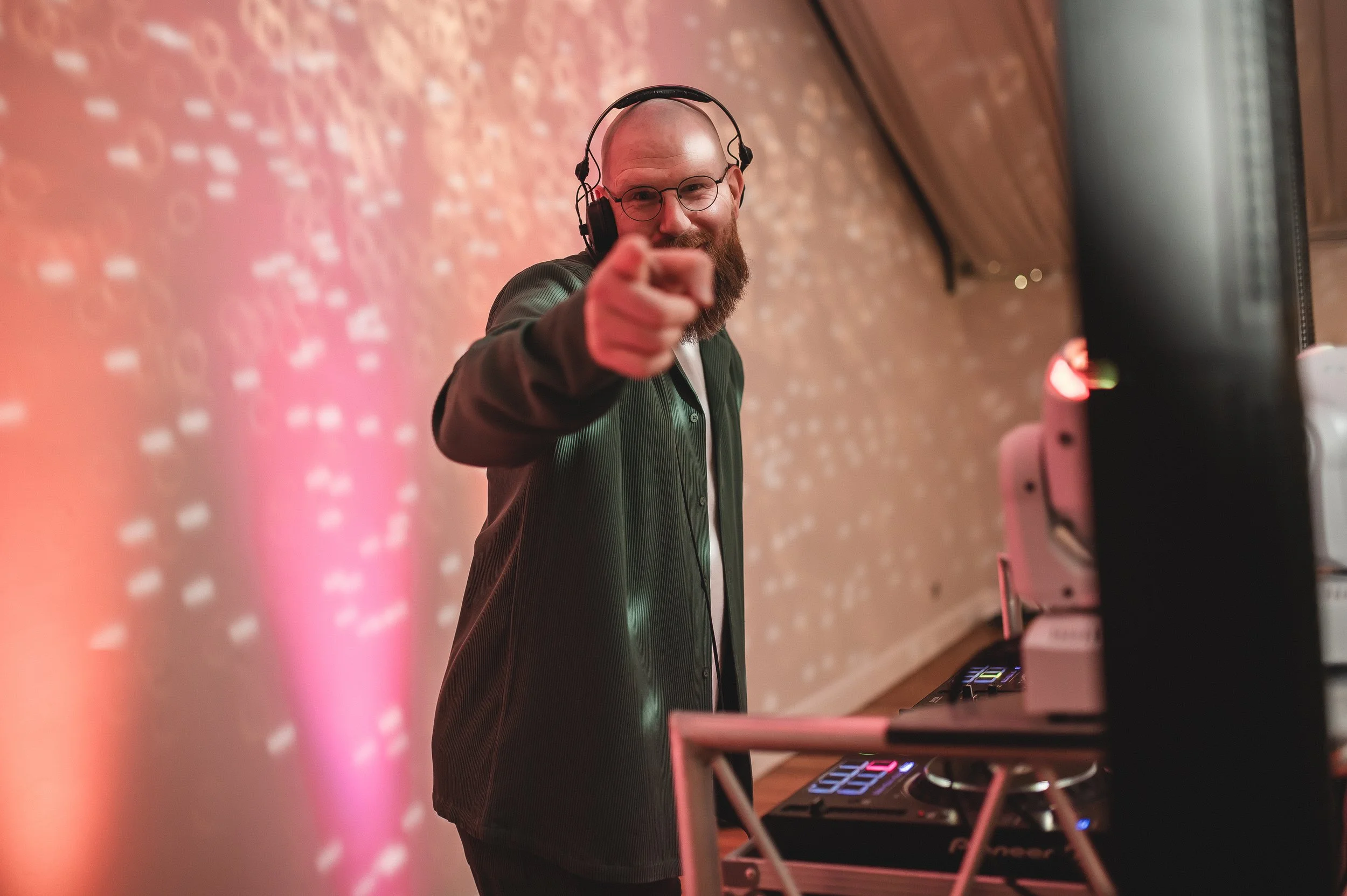 A Married to the Music wedding DJ performs live, creating an unforgettable party atmosphere with professional lighting and sound. The DJ stands behind the decks, pointing toward the crowd, ready to bring the dance floor to life with expertly mixed tr