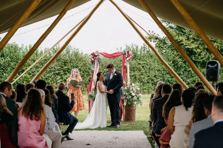 A joyful outdoor wedding ceremony under a tipi, featuring a bride and groom standing together beneath a floral archway with red and pink blooms, as a smiling Frances Celebrates You in a vibrant orange floral dress leads the vows. Guests watch from wo