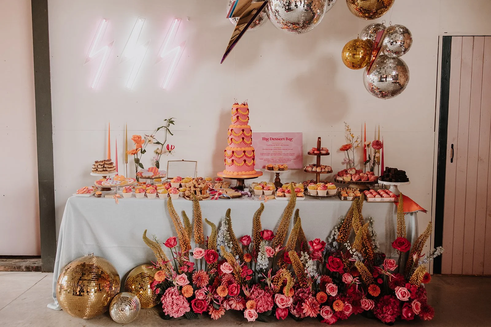 A vibrant dessert table setup at The Giraffe Shed wedding venue in Wales, styled with bold retro details and modern pops of colour. The table features a statement pink and orange tiered wedding cake surrounded by cupcakes, brownies, and sweet treats,
