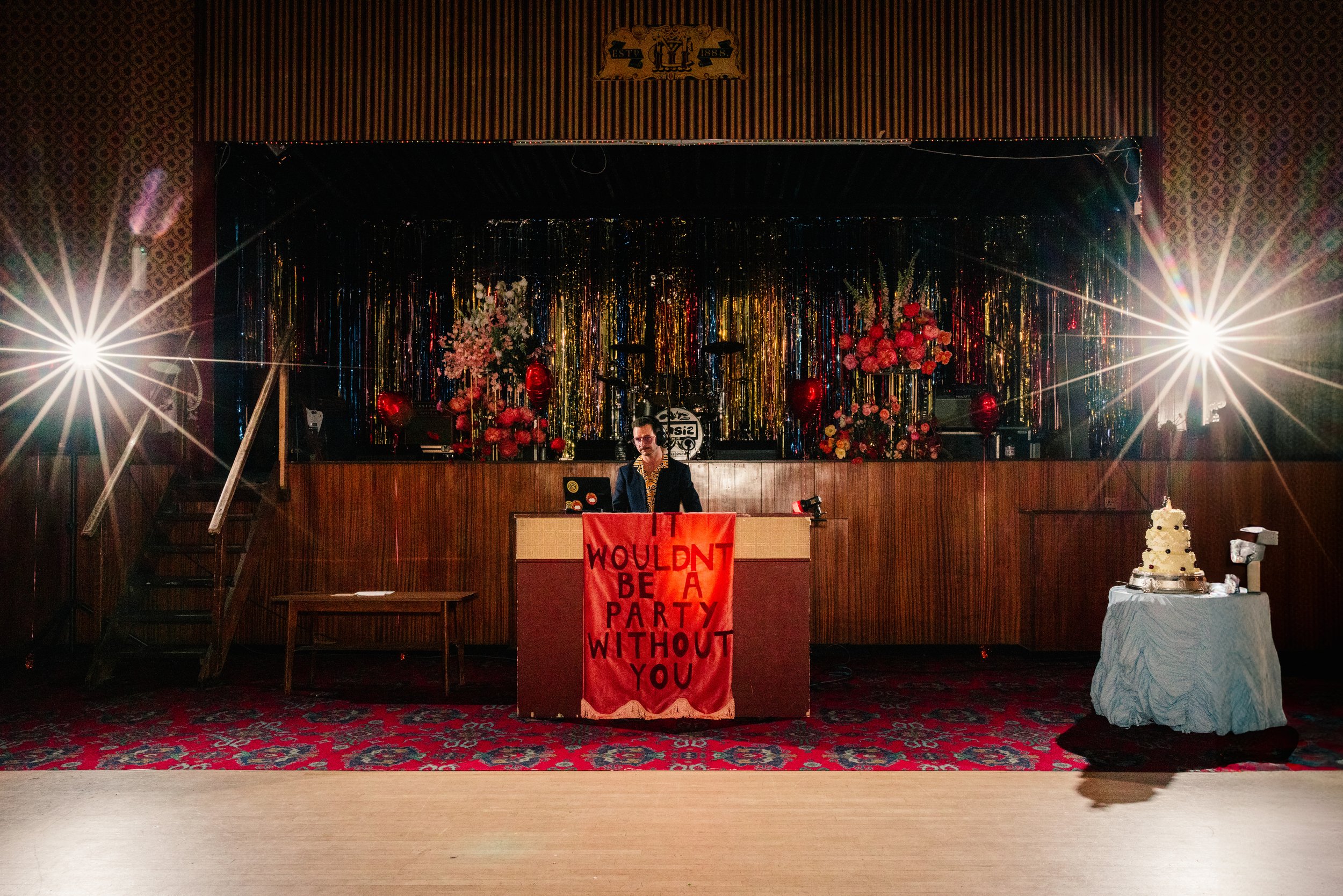 A wide-angle wedding reception photograph taken at the Mildmay Club captures a dramatic, retro-styled stage setup with bold lighting and vibrant décor. In the center, a DJ stands behind a wooden booth draped with a red banner reading “Wouldn’t Be a P