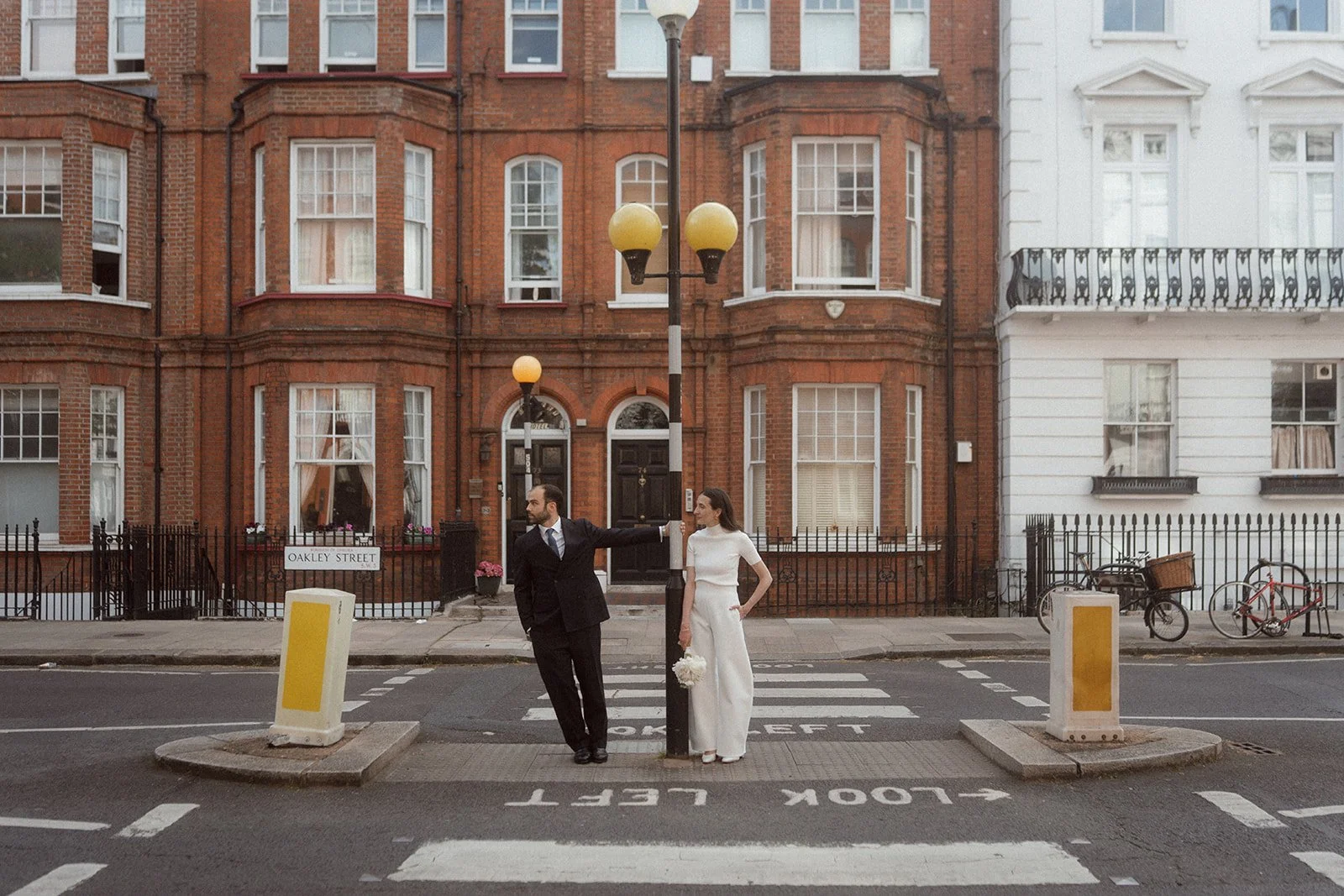 A couple poses at a pedestrian crossing on a quiet London street with red brick townhouses in the background, with the groom leaning against a lamppost in a dark suit and the bride in a white jumpsuit holding a bouquet.