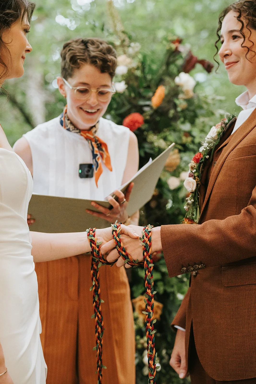 A beautiful outdoor wedding ceremony featuring a couple during a symbolic hand-fasting ritual, led by wedding celebrant Chloe Green. The couple’s hands are gently tied together with colourful braided cords, symbolising unity and commitment, surrounde