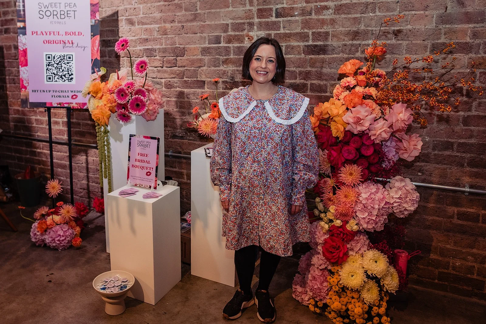 A wedding florist in a floral dress is smiling next to her display at a wedding fair.