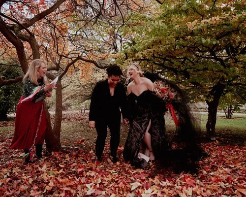 Outdoor autumn wedding ceremony with a joyful same-sex couple laughing among golden leaves. The wedding celebrant, holding a folder, officiates beneath colourful fall trees as the brides - one in a black gown and the other in a dark suit - celebrate 
