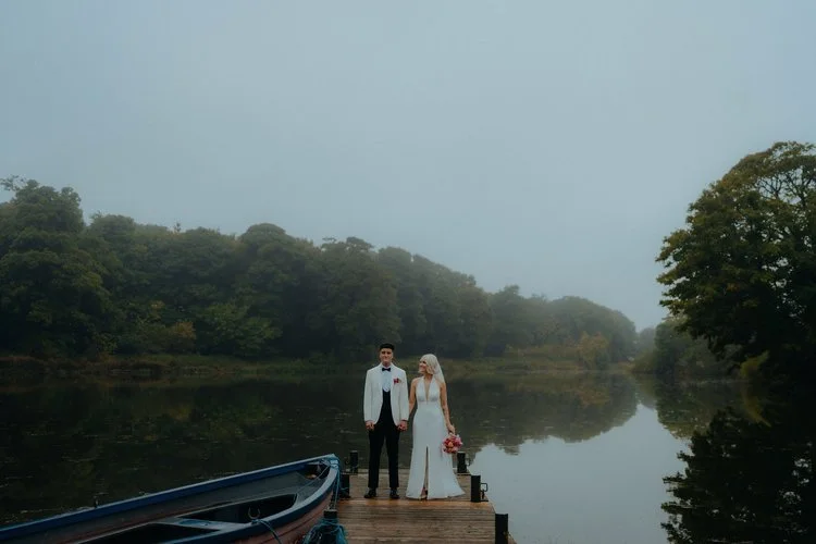 A beautifully moody scene captured by F.D Young Photography: a couple walking hand in hand along a quiet coastal path, sharing a clear umbrella as the sky stretches endlessly above them. The muted tones, open landscape, and distant lighthouse create 