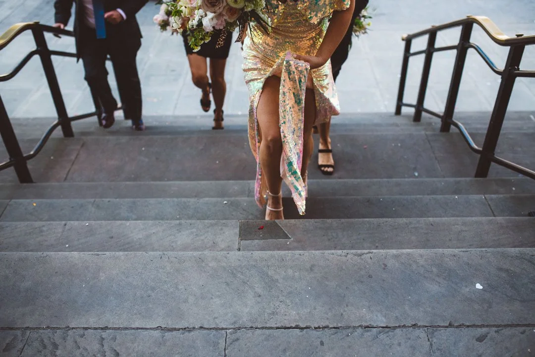 A dynamic and stylish wedding moment captured by Barbara K Photography, showing a bride in a shimmering sequin gown climbing a set of stone steps while holding a bouquet of soft pastel flowers. The perspective focuses on the movement of her dress and
