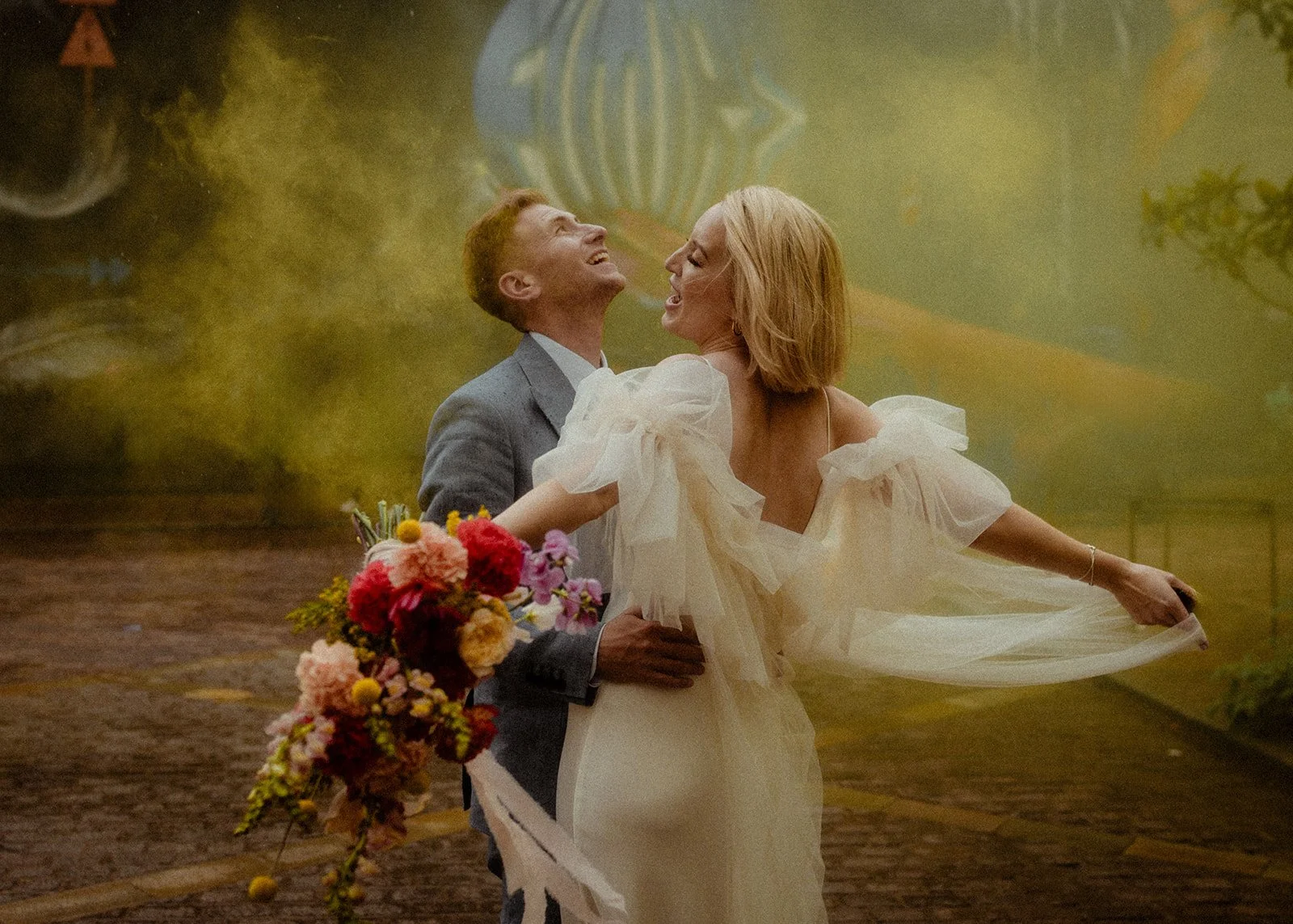 A couple stands wrapped in a moment of vivid celebration as coloured smoke drifts around them, with the groom holding the bride at the waist while she joyfully lifts her sheer, ruffled sleeves and leans back into the scene, her bouquet of bold pink, 
