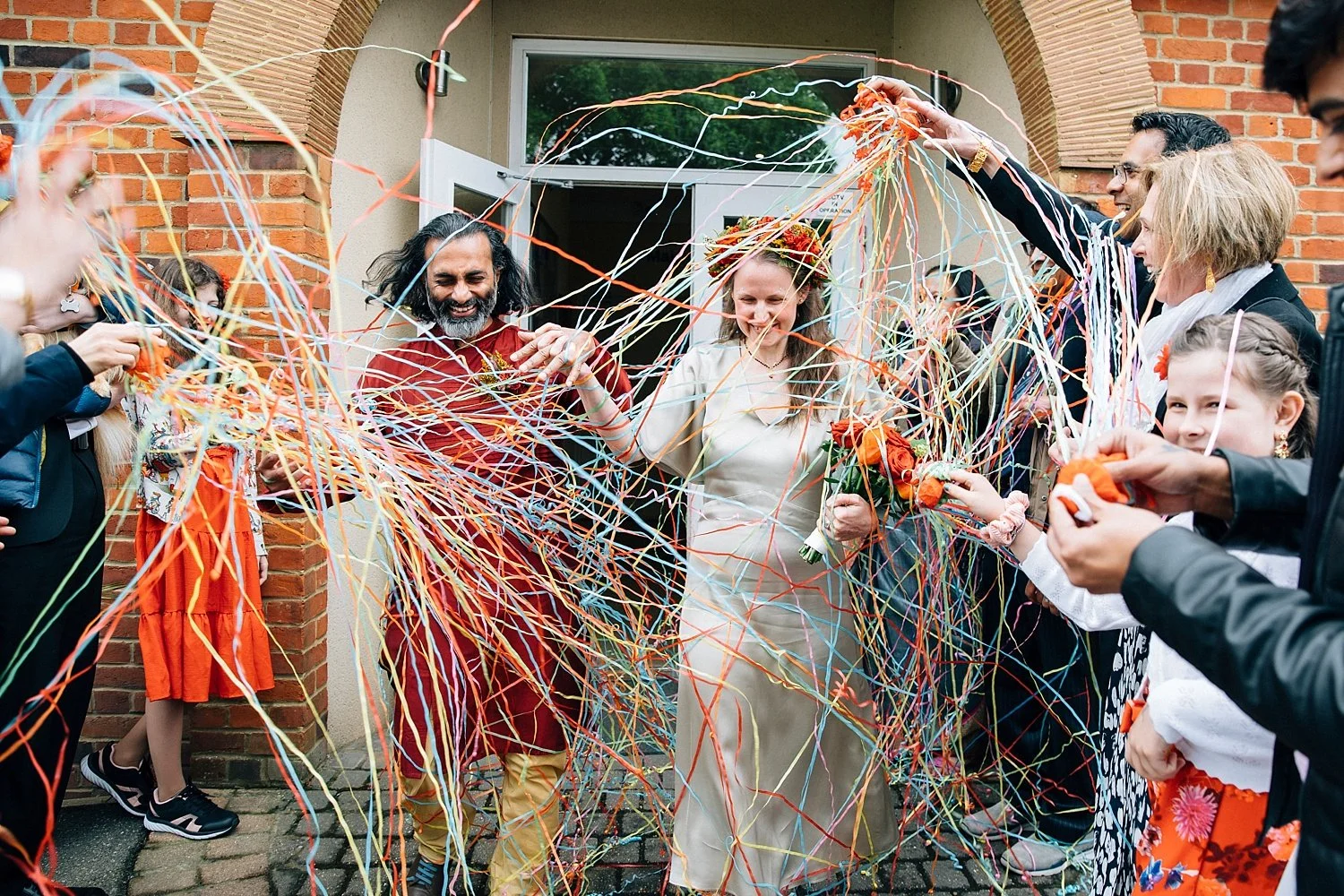 A joyful wedding exit captured by Paola de Paola Photography shows a newly-married couple walking through a vibrant shower of rainbow-coloured streamers outside their ceremony venue. Friends and family form a lively guard of honour, filling the air w