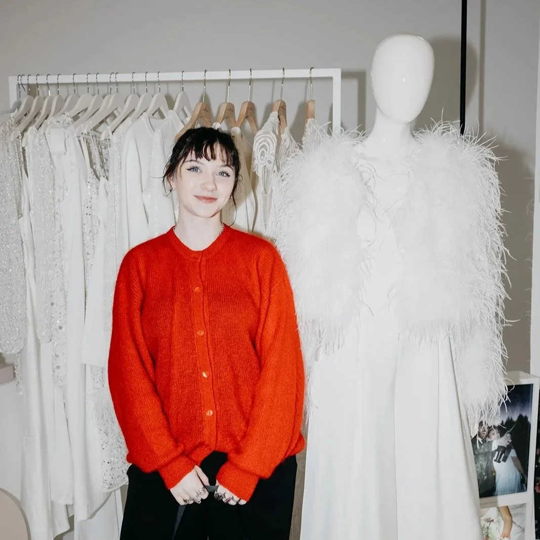 A woman in a red cardigan is standing in front of some modern bridal outfits at an MNT wedding fair.