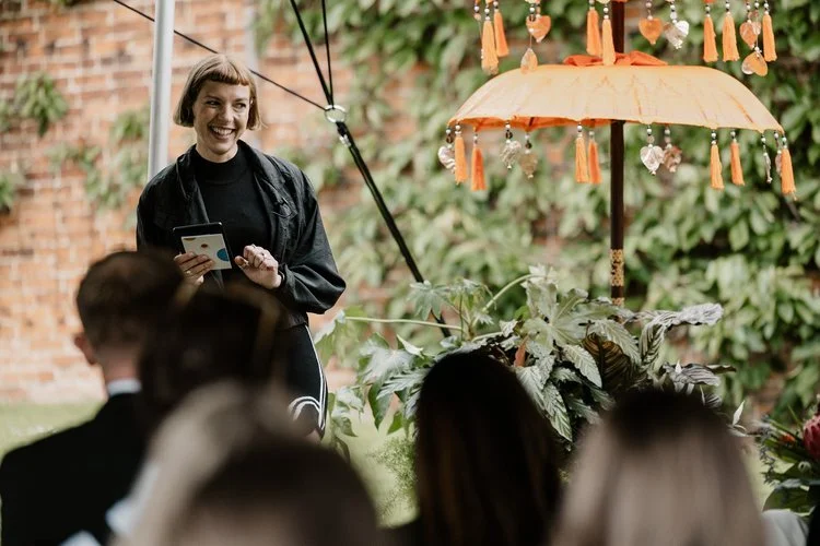 Modern wedding celebrant Tab Taylor from Say it Right Ceremonies leads an intimate outdoor ceremony beneath a vibrant orange parasol adorned with tassels and crystals. Speaking warmly to seated guests, the celebrant’s confident and joyful presence cr