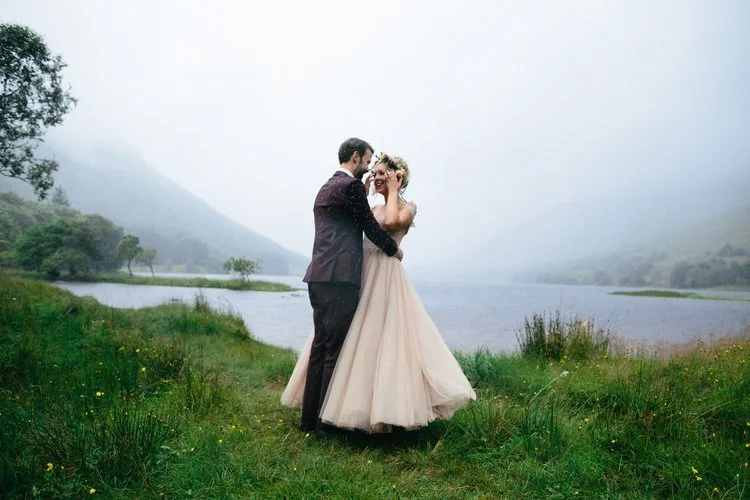 A dreamy outdoor wedding portrait captured by Mirrorbox Photography, featuring a couple embracing beside a misty loch in the Scottish Highlands. The bride’s soft, flowing blush-toned gown moves gently in the breeze, complemented by her floral crown, 