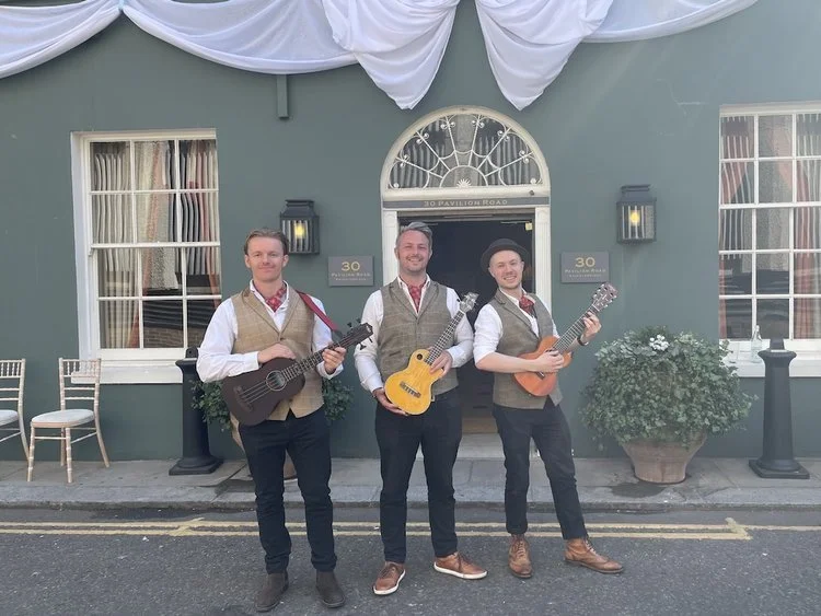 A lively wedding band, The Ukes of Hazard, pose with their ukuleles outside a stylish London wedding venue. The three musicians are dressed in coordinated rustic-chic outfits featuring beige waistcoats, white shirts, and red patterned neckerchiefs, p