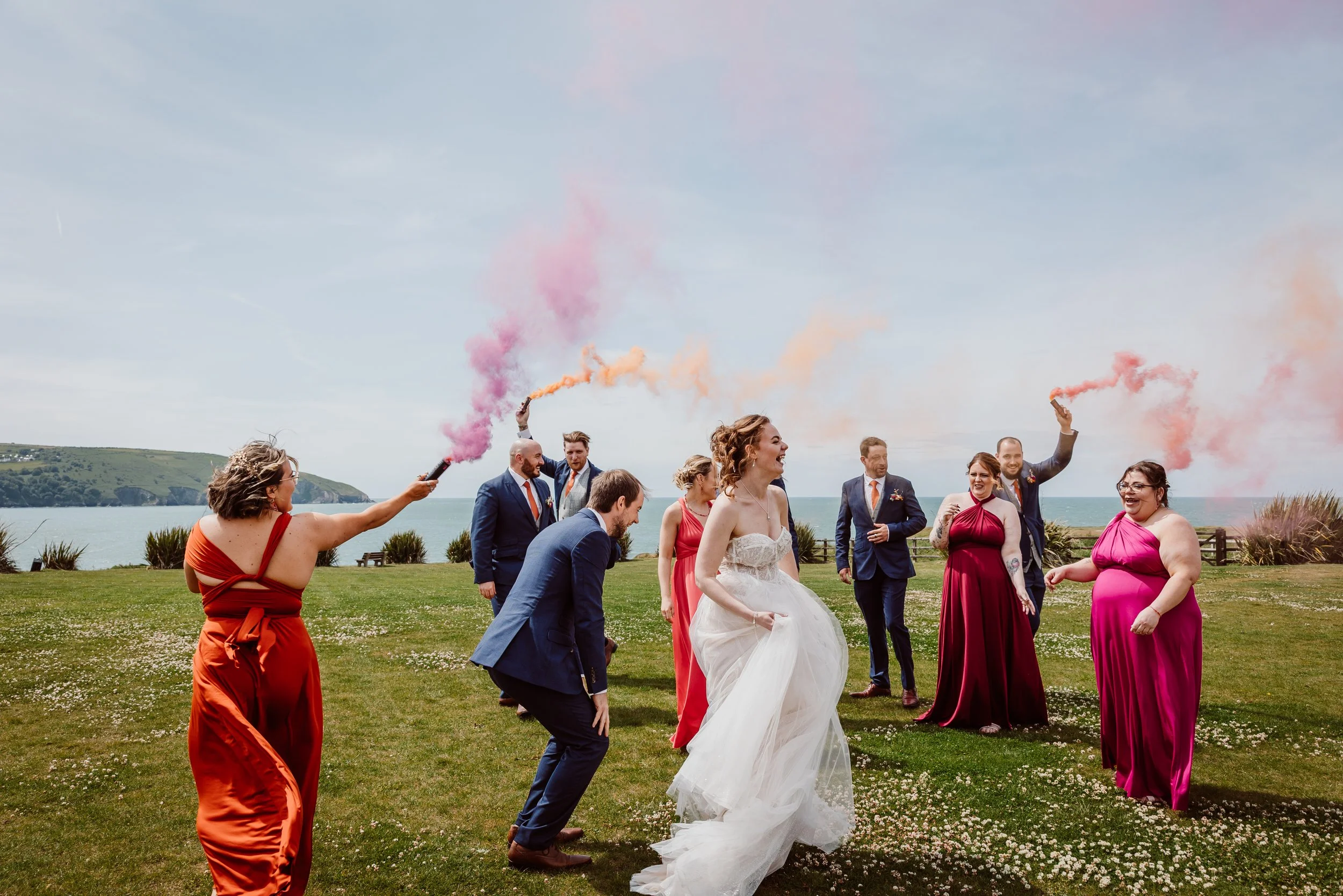 A fun and vibrant wedding party moment captured by Joy Louise Photography, showing the bride and her friends celebrating outdoors with colourful smoke flares. Set against a stunning coastal backdrop with blue skies and rolling green hills, the scene 