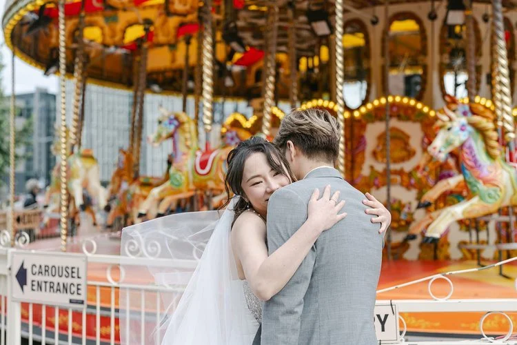 A joyful bride and groom share a warm, intimate embrace beside a vibrant vintage carousel, beautifully captured by Christy Photography. The colourful fairground backdrop adds energy, movement and personality to the scene, highlighting the couple’s na