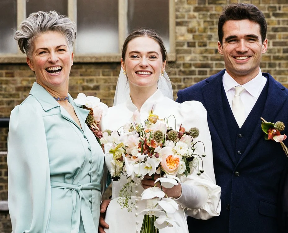 Arabella stands alongside the couple for a posed portrait, all three facing the camera and smiling. The bride holds a large bouquet of mixed flowers, while the celebrant and partner stand close on either side, framed by a brick wall backdrop in natur