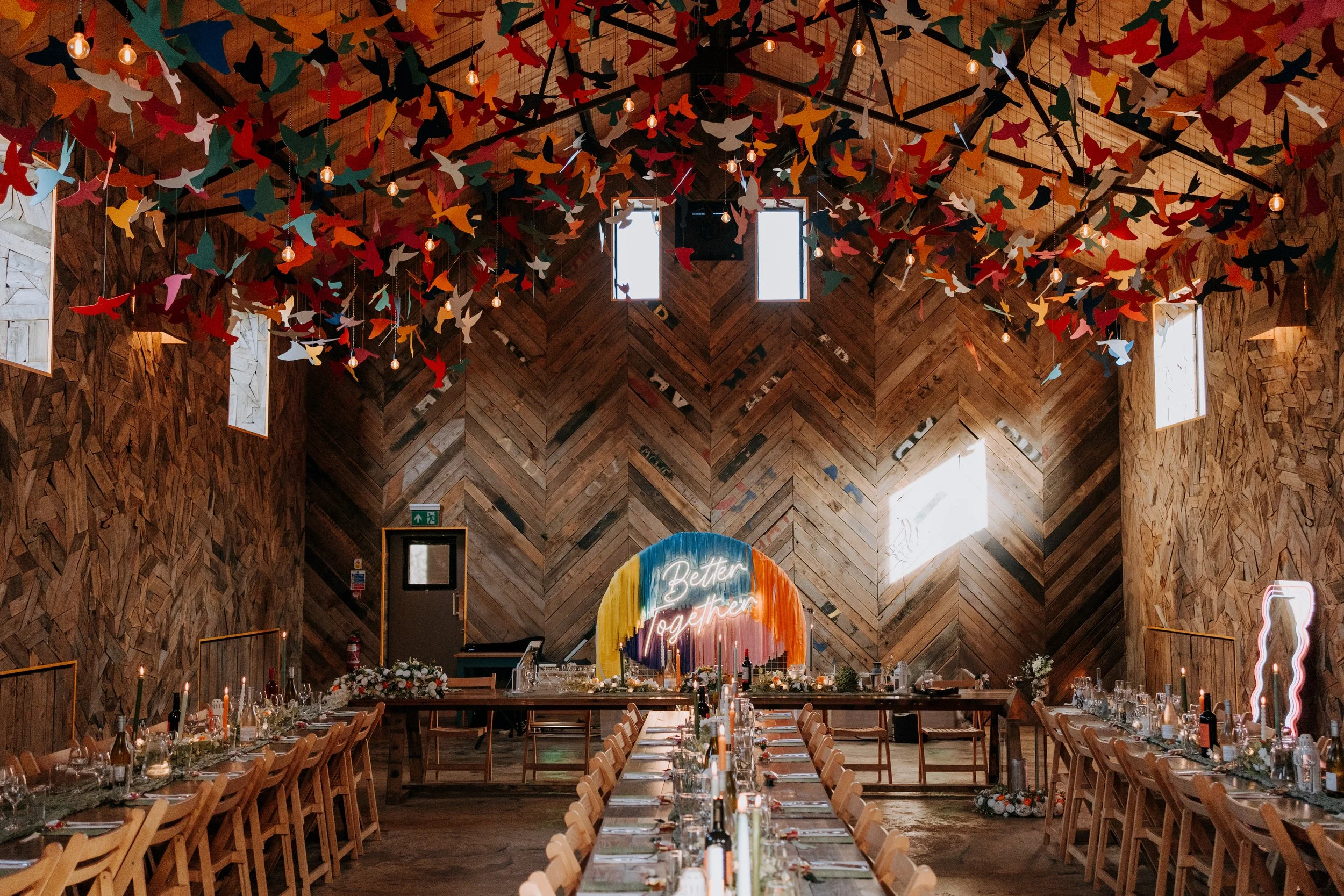 Long wooden tables set for dinner stretch down a high ceilinged hall with warm timber walls arranged in a herringbone pattern, while hundreds of brightly coloured hanging birds cluster overhead beside exposed bulbs and natural light pours through tal