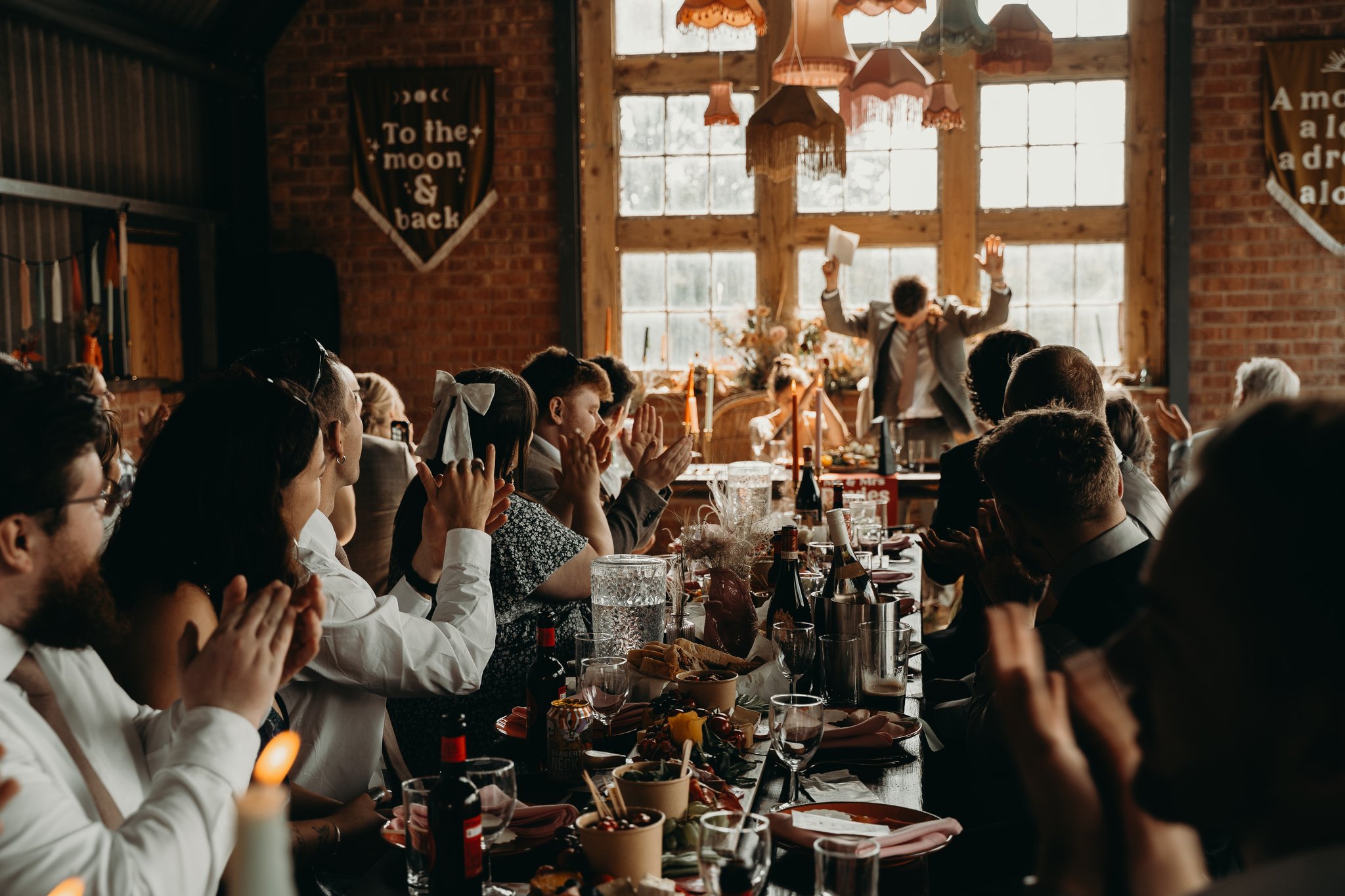A joyful wedding reception inside The Giraffe Shed in Wales, capturing guests clapping and cheering during heartfelt speeches beneath the venue’s signature industrial beams and large rustic windows. The long banquet tables are styled with modern boho