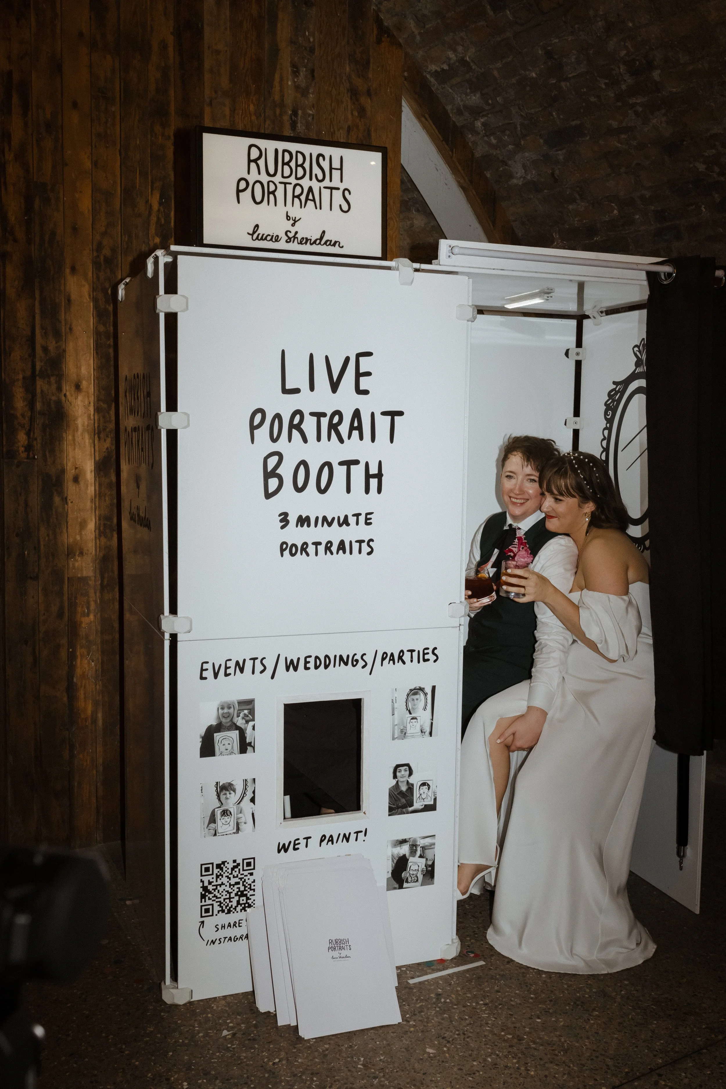 A joyful couple sharing drinks inside the ‘Rubbish Portraits’ live portrait booth by Lucie Sheridan. The booth offers quick 3-minute portraits for events, weddings, and parties, featuring a fun hand-drawn design with photo samples and a ‘Wet Paint!’ 