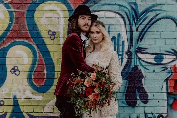 Bohemian wedding couple captured against a colourful graffiti wall in Liverpool, featuring the bride with soft natural makeup by Glamorama Make-up Artist Liverpool. A unique urban wedding look blending artistic street style with romantic bridal beaut
