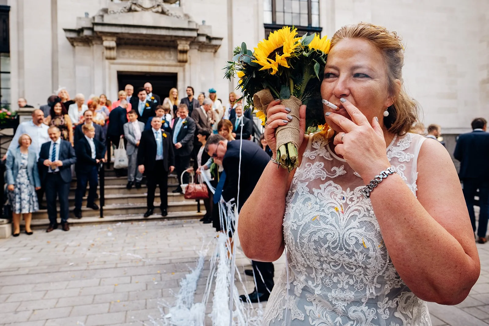 A woman in an ornate lace dress is pictured standing in the foreground outside a UK registry office, holding a sunflower bouquet in one hand and a cigarette in the other, while a large group of formally dressed guests gathers on the steps behind her.