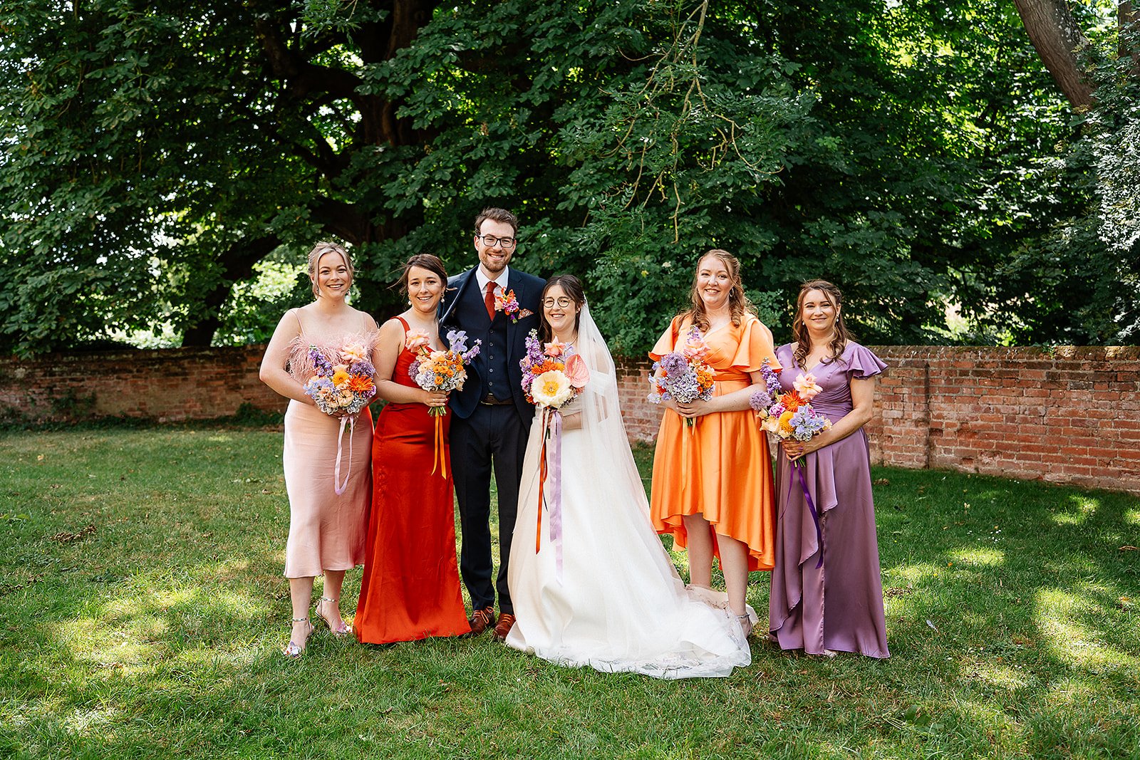 Colourful summer wedding portrait captured by Ben Cotterill Photography, featuring the bride and groom with bridesmaids in pink, red, orange, and purple dresses. Each holds a vibrant bouquet by Sweet Pea Sorbet Floristry with seasonal blooms in coord