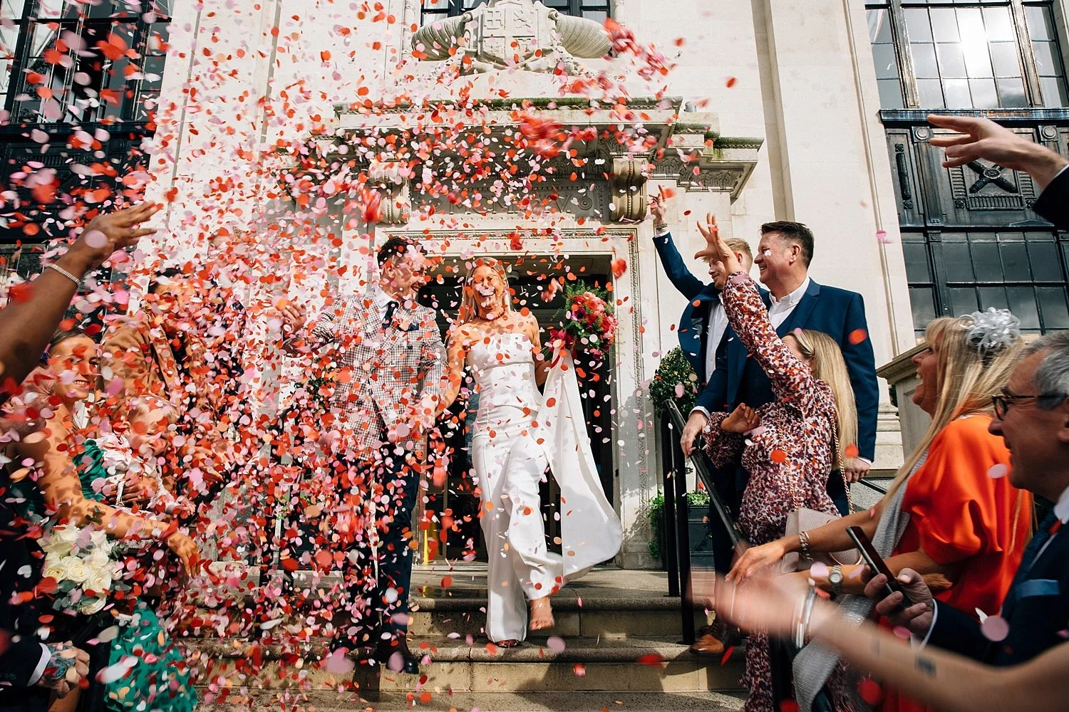  A burst of joyful chaos as a couple steps out into a storm of pink and red confetti, completely engulfed in it. They’re laughing, holding hands, and moving down the steps together with infectious energy. The bride wears a sleek white jumpsuit with a