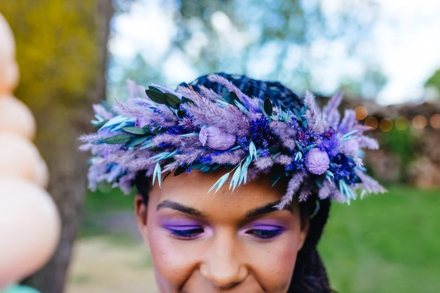 A striking dried-flower crown in electric purples and blues sits proudly atop the model’s head, its mix of fluffy grasses, painted leaves and bold botanicals echoing the vivid violet eye makeup beneath, creating a dreamy, otherworldly look against th