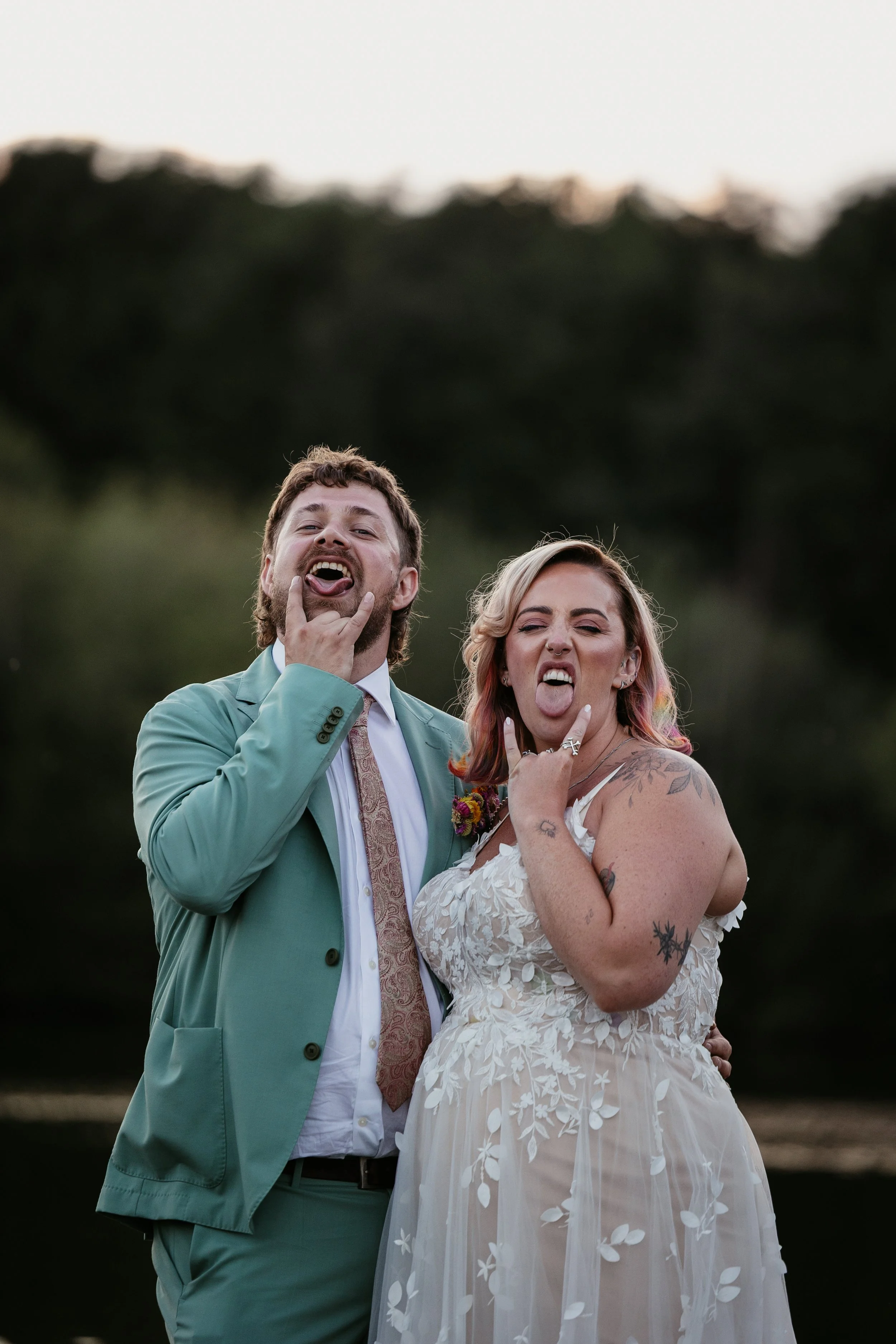 A fun and personality filled portrait captured by Joy Louise Photography, showing a newly married couple pulling playful faces at the camera during golden hour. The groom stands confidently in his pastel green suit while the bride glows in a floral l