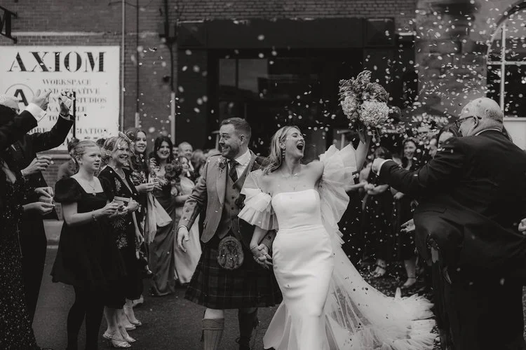 A couple walk hand in hand through a lively confetti tunnel surrounded by cheering guests, captured with striking emotion and atmosphere by F.D Young Photography. The bride’s dramatic ruffled sleeves and the groom’s traditional kilt contrast beautifu