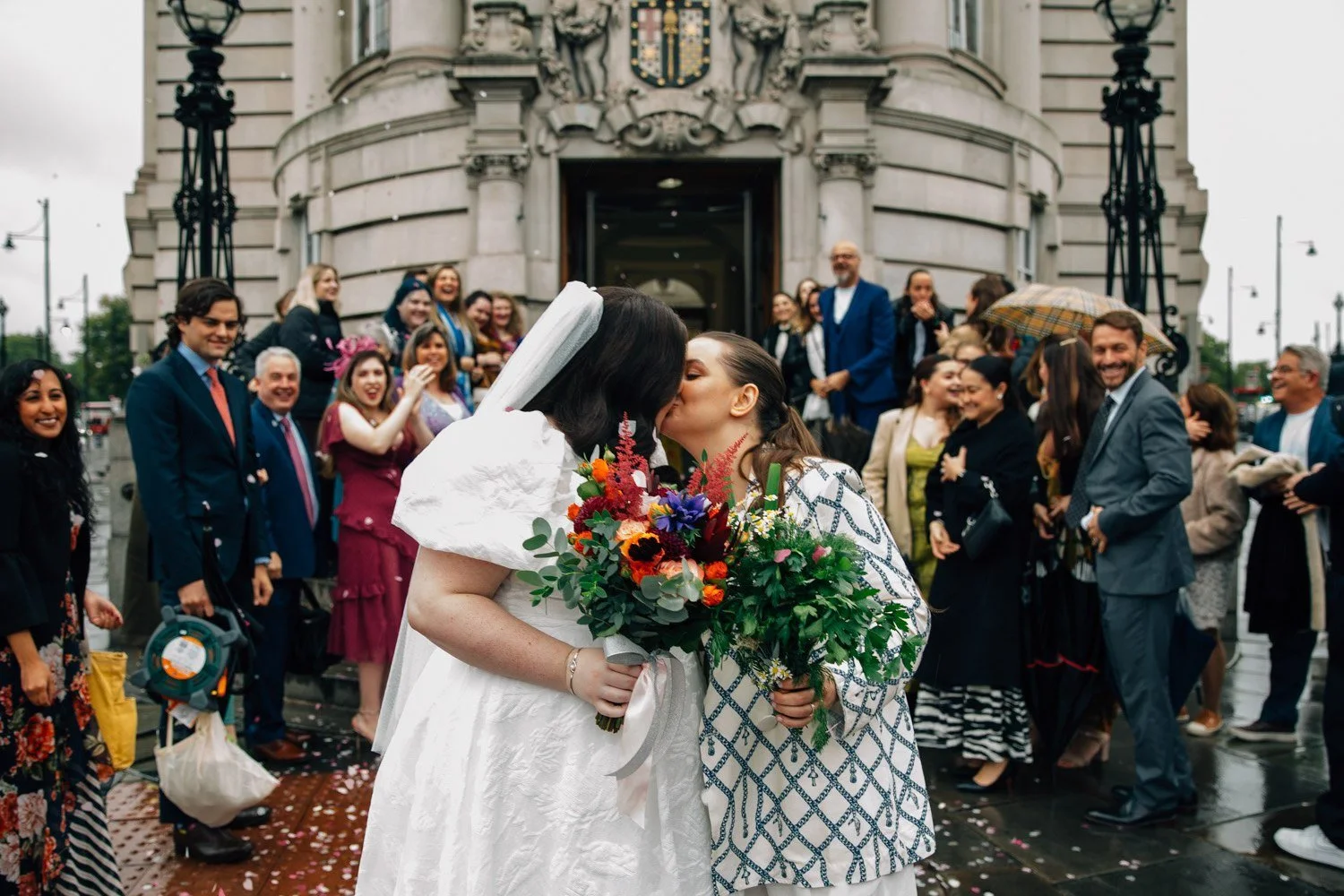 A joyful same-sex wedding celebration outside a grand city hall, featuring two brides sharing a kiss while holding vibrant, colourful bouquets. Guests surround them cheering and throwing confetti, creating a lively, candid moment full of love and cel