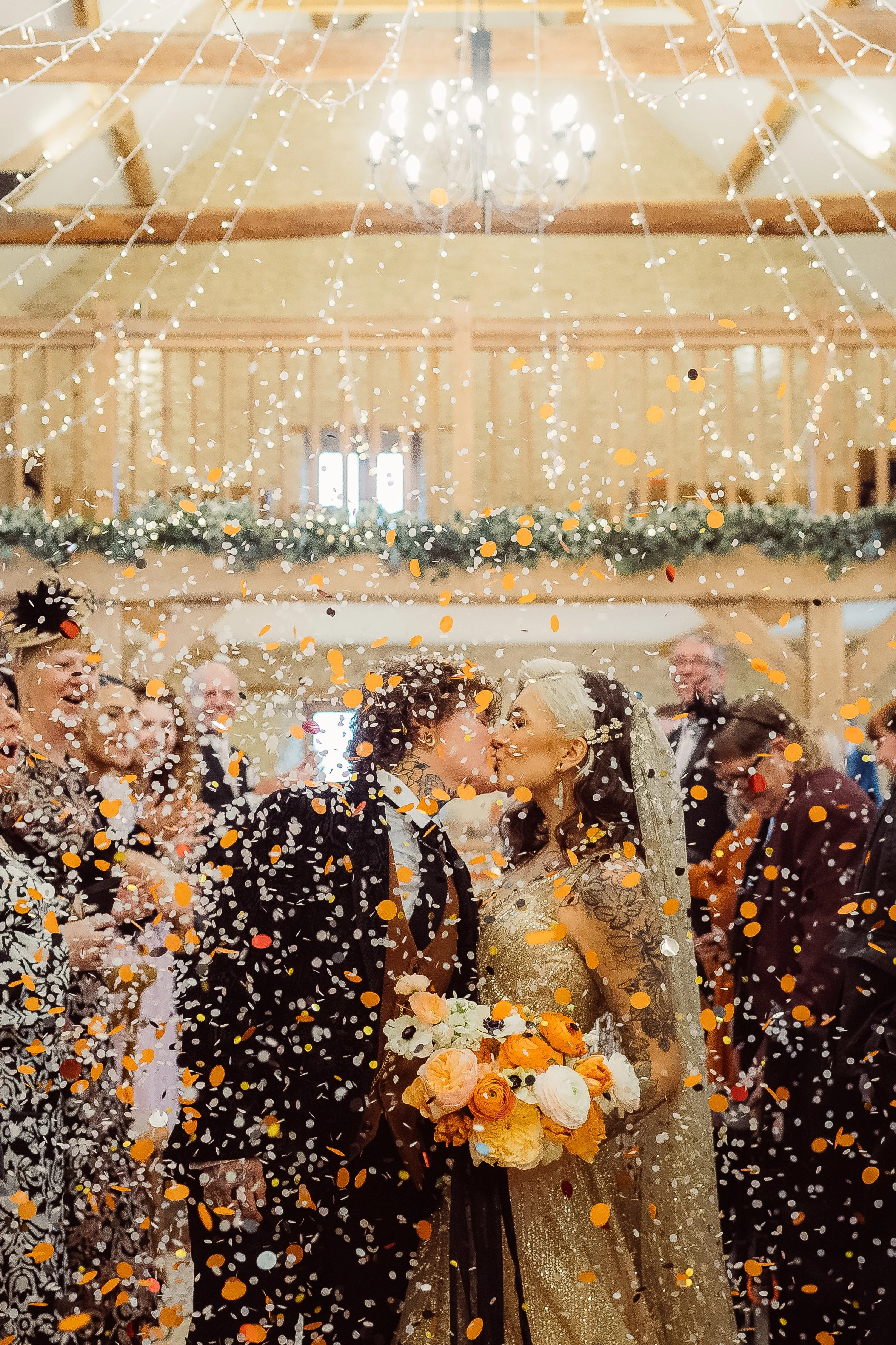 A vibrant confetti-filled wedding moment captured by Gemma Gaskins Photography, showing a newlywed couple sharing a kiss beneath twinkling fairy lights. The warm indoor barn setting, colourful falling confetti, and bold bouquet create a joyful, cinem