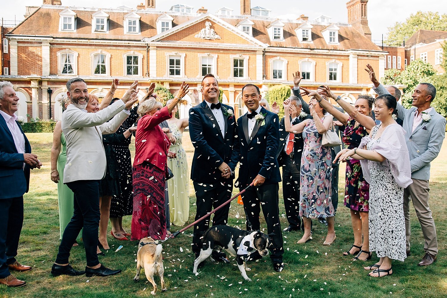 A wave of pure celebration surrounds two grooms as they stand beaming in front of a grand historic building, hands clasped and hearts wide open. Their guests form a joyful semicircle around them, tossing soft white petals into the warm summer air. Th