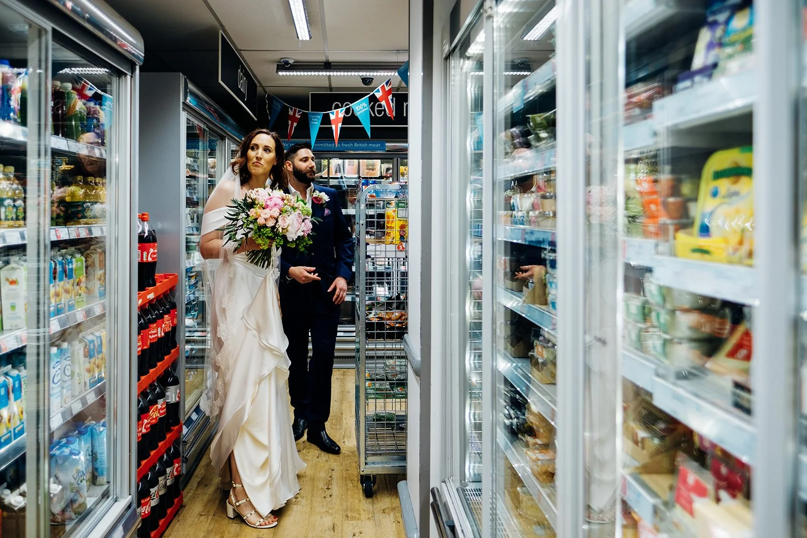 A couple dressed in formal wedding attire stands together in the chilled aisle of a small UK convenience store, with the woman holding a bouquet of pink and green flowers and the man in a dark suit beside her. They are surrounded by refrigerated shel